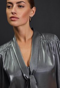 Bluză gri mătăsoasă cu guler legat, având mâneci plisate și o textură netedă. Design minimalistic cu un decolteu adânc în formă de V.