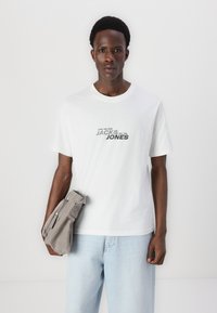 Camiseta blanca de algodón con texto gráfico negro, corte relajado. Pantalones de mezclilla azul claro, bolsa de tela gris apretada bajo el brazo derecho.