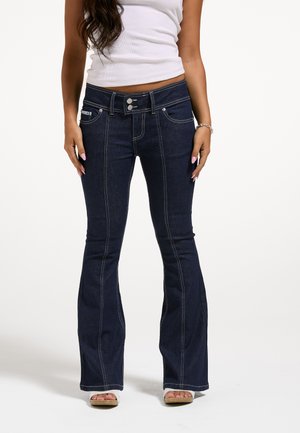 Femme portant un jean évasé bleu foncé avec des détails de couture à l'avant et une fermeture double bouton, associé à des sandales blanches et un top blanc sans manches.