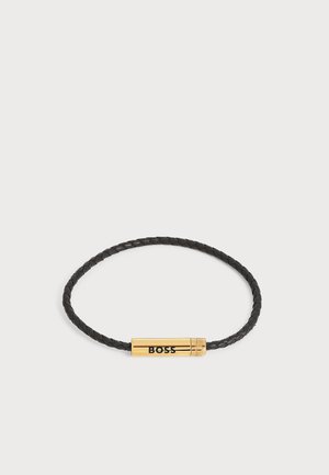 Sort flettet læderarmbånd med en guldcylindrisk lukning graveret med ordet "BOSS" i sorte bogstaver.