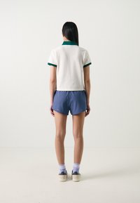 Polo branca de mangas curtas com um colarinho verde, combinada com calções azuis. O modelo usa meias roxas e sapatos bege com detalhes escuros.