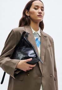 Μαύρη δερμάτινη τσάντα με τριγωνικό σχήμα, με φερμουάρ και υφή, κρατημένη μπροστά από ένα καφέ υπερμεγέθες σακάκι.