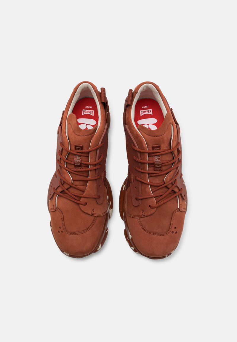 Brązowe zamszowe trampki z zaokrąglonymi noskami, wyraźnymi sznurówkami, wyściełanymi kołnierzami i teksturowanymi gumowymi podeszwami z charakterystycznym wzorem.