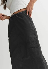 Vrouw die de trekkoord aanpast van een zwarte cargorok met klepzak, terwijl ze een witte geribde crop top draagt tegen een effen achtergrond.