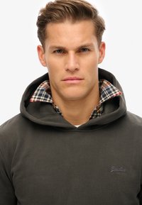 Sudadera gris oscuro con una gran capucha y forro de cuadros visible en el cuello. Presenta una sutil marca en el pecho. Textura suave.