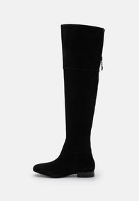 Botas negras de ante hasta la rodilla, con un diseño ajustado, punta afilada y cremallera lateral para mayor facilidad de uso. Tacón plano.