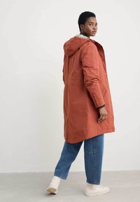 Lång orange regnjacka med huva, tryckknappsstängning, sidofickor och fodrad krage, bärs över blå vida jeans och vita skor.