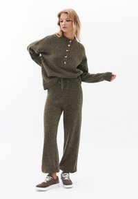 Olivengrüner Strickpullover mit gerippter Struktur und Knopfleiste, kombiniert mit passender lockerer Hose und braunen Wildledersneakern.