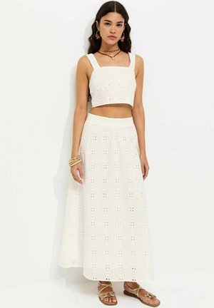 Młoda kobieta ubrana w biały ażurowy crop top i pasującą długą spódnicę z złotymi sandałami i biżuterią na jednolitym tle.