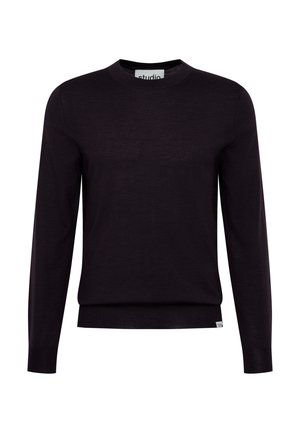 Ung mand med kort krøllet hår iført en mørkelilla langærmet skjorte og sorte bukser, stående mod en lys væg med hænderne i lommerne.