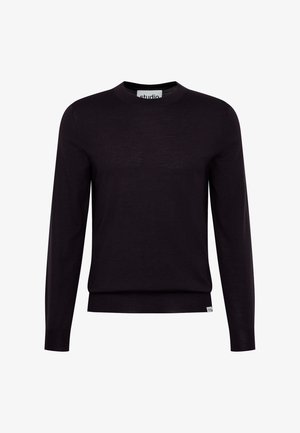 Junger Mann mit kurzen lockigen Haaren, der ein dunkelviolettes Langarmshirt und schwarze Hose trägt, steht mit den Händen in den Taschen vor einer hellen Wand.