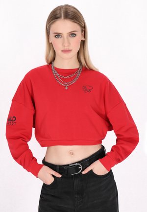 Młoda kobieta o blond włosach nosi czerwony krótki sweter z łańcuchami oraz czarne spodnie z wysokim stanem i paskiem, ręce w kieszeniach.