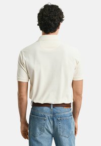 Beige pikétröja med kort ärm, slät textur och krage, tillsammans med ljusblå denimjeans med bakfickor och ett brunt bälte.