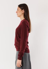 Maglione in lana bordeaux con maniche lunghe, scollo a girocollo e polsini a costine. Indossato con una gonna grigia, mostrato in vista laterale.
