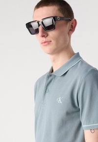 Sorte solbriller med firkantede rammer og mørke linser. Lyseblå polo-skjorte med teksturert stoff og en liten, hvit logo på brystet.