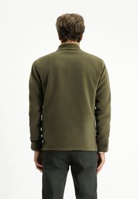 Giacca in pile verde oliva con colletto alto, maniche lunghe e una trama liscia. Indossata con pantaloni verde scuro. Design semplice, senza motivi visibili.