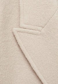 Revers et rabat de poche d'un manteau en laine beige avec un tissu doux et texturé et des détails de couture nets et structurés.