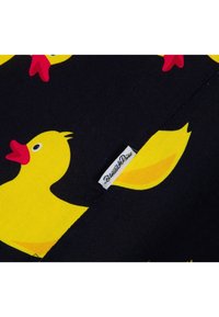 Gelber Gummiente-Muster auf schwarzem Stoff mit einem kleinen weißen Etikett, auf dem "Blumat & Co" in schwarzem Text steht. Glatte Textur, verspieltes Design.