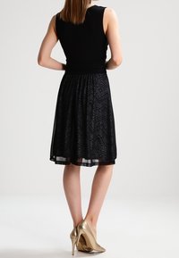 Musta hihaton mekko, jossa on istuva yläosa ja levenevä hame. Hameessa on viistokuvio. Yhdistetty metallinhohtosiin kultaisiin korkokenkiin.