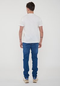T-shirt bianca in cotone con maniche corte, vista da dietro, abbinata a jeans in denim blu e scarpe da ginnastica di colore chiaro.