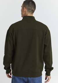 Mand med kort hår og et lille ar på hovedbunden iført en mørkegrøn sweatshirt og blå jeans, der vender væk fra kameraet.
