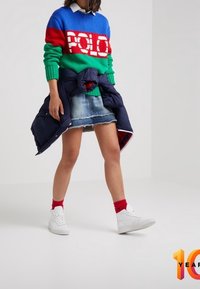 Person med en färgglad tröja med "POLO"-text, denimkjol, röda strumpor, vita sneakers och en marinblå jacka knuten runt midjan.