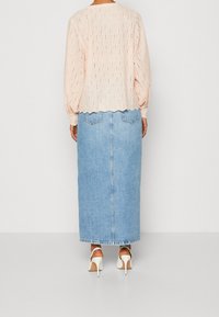 Femme portant un chemisier à manches longues en œillet rose clair, une jupe midi en denim bleu clair et des sandales blanches à talons hauts, debout de dos.