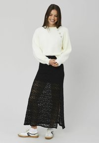 Pull blanc et moelleux à col rond avec trois accents décoratifs, associé à une jupe longue noire en dentelle avec une fente latérale et un détail texturé.