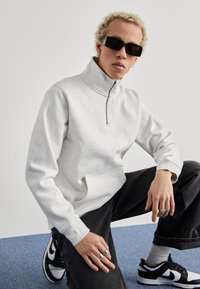 Grå zip-neck sweatshirt med en frontlomme, parret med sorte denimbukser og sort-hvide sneakers. Har en afslappet pasform og ribbede manchetter.