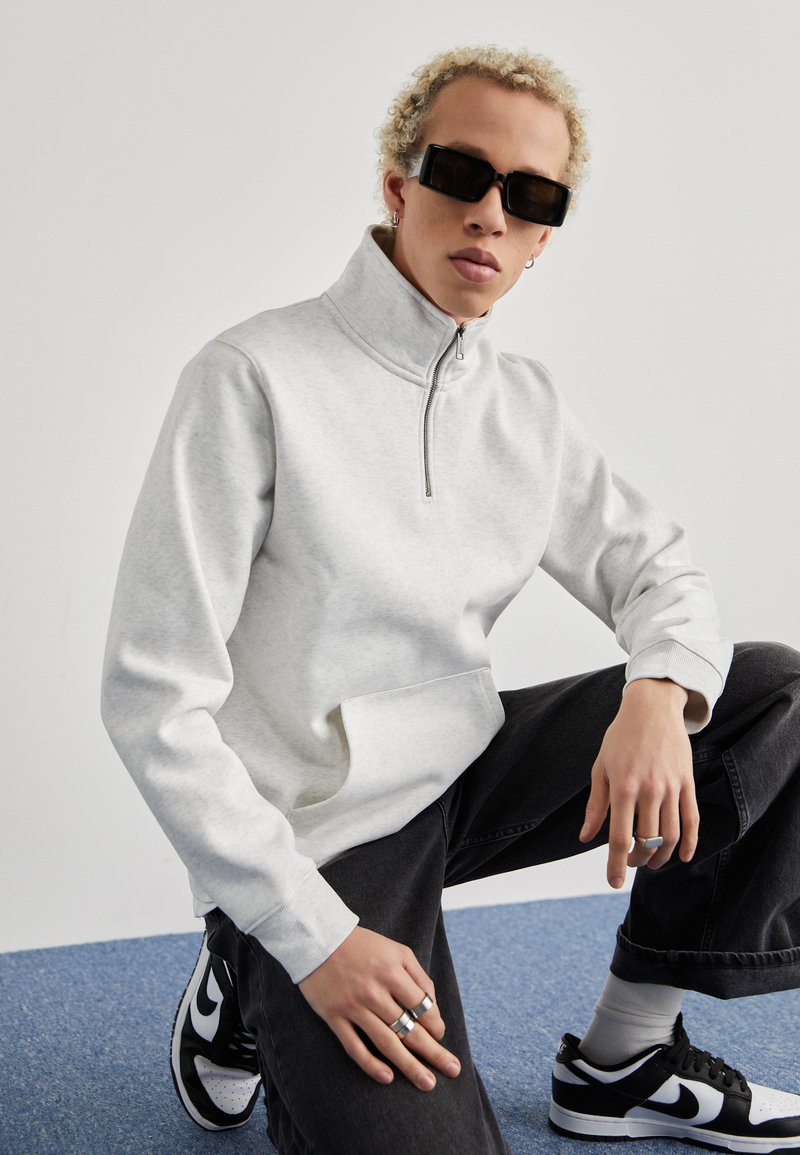Grå zip-neck sweatshirt med en frontlomme, parret med sorte denimbukser og sort-hvide sneakers. Har en afslappet pasform og ribbede manchetter.