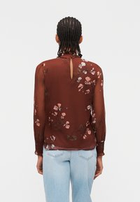 Personne aux cheveux tressés portant un chemisier marron transparent à motifs floraux avec une ouverture en forme de clé à l'arrière, et un jean taille haute bleu clair.