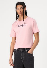 Šviesiai rožinė medvilninė marškinė su tamsiai mėlynu "Pepe Jeans" logo centre, derinama su tamsiai pilkais džinsais. Trumpi rankovės ir standartinis fasonas.