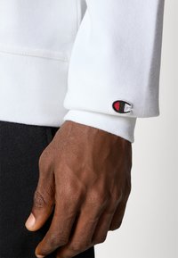 Sweatshirt blanc avec des poignets côtelés, arborant un petit logo brodé avec des accents rouges et noirs sur la manche. Du tissu sombre est visible en dessous.