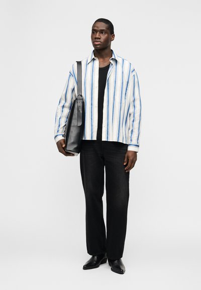 Άνδρας που φοράει μια λευκή και μπλε ριγέ πουκαμίσα πάνω από μαύρο μπλουζάκι, μαύρο παντελόνι, μαύρα παπούτσια, κρατώντας μια μεγάλη μαύρη τσάντα ώμου, στέκεται μπροστά σε λευκό φόντο.