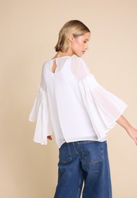 Hvit bluse med gjennomsiktige, klokkeformede ermer og detalj bak med nøkkelhull. Kombinert med denim culottes. Glatt tekstur, moderne design.