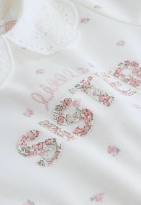 Fehér ruha virágos hímzéssel. "Kis húg" felirat mintás anyagon. Csipkés gallérral és apró rózsaszín virágos díszítéssel.