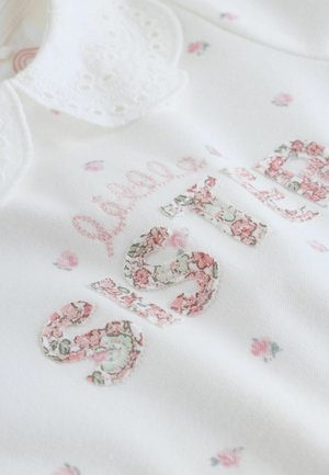 Biely odev s kvetinovou výšivkou. Text „Malá sestra“ na vzorovanej tkanine. Má čipkový golier a malé ružové kvetinové ozdoby.
