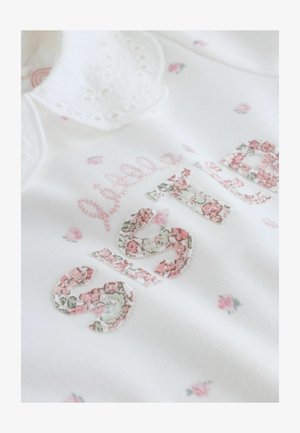 Vêtement blanc avec broderie florale. Texte « Petite Sœur » en tissu à motifs. Présente un col en dentelle et de petits accents floraux roses.