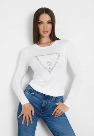 Maglietta bianca a maniche lunghe con logo "GUESS" in argento decorato a forma di triangolo e punto interrogativo. Abbinata a jeans blu a vita alta.