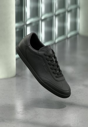 Baskets en cuir noir avec lacets, marquage "Guess" sur le côté et la languette, flottant au-dessus d'une surface grise réfléchissante près d'un mur en blocs de verre.