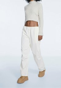 Cremfarbener, gerippter Cropped-Pullover mit hohem Kragen, kombiniert mit weißen, elastischen Hosen und beigen, klobigen Plateau-Schuhen. Glatte Texturen.