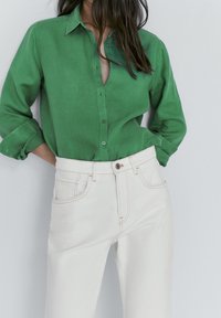 Woman wearing a green buttoned shirt with rolled sleeves tucked into high-waisted white jeans, standing with hands behind her back.