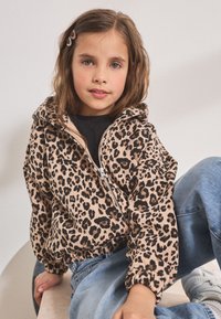 Jachetă de fleece cu imprimeu leopard, cu fermoar, glugă și manșete elastice, asortată cu blugi de culoare albastru deschis și pantofi sport albi. Fată așezată pe o suprafață.