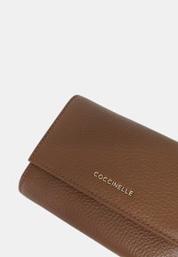 Brown leather wallet features a textured surface, a flap closure, and embossed “Coccinelle” logo on the front. Slim and rectangular design.