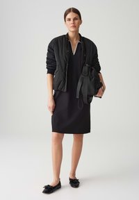 Schwarze gesteppte Bomberjacke über einem schwarzen Kleid mit Kragen, kombiniert mit schwarzen Ballerinas mit Schleifen. Schwarze Handtasche mit Schulterriemen.