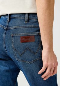 Los jeans de mezclilla azul presentan un acabado texturizado, un diseño de corte recto y una parche de cuero marrón en el bolsillo trasero con un patrón de costura en zigzag.