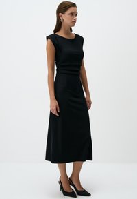 Abito midi nero con spalle strutturate, tessuto liscio e corpetto aderente. Abbinato a décolleté nere con cinturino. Design semplice ed elegante.