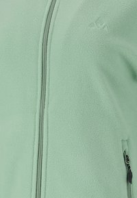 Helle grüne Fleecejacke mit durchgehendem Reißverschluss, strukturiertem Stoff und einer Seitentasche mit schwarzem Reißverschluss.