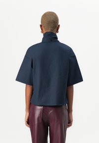 Kortsleeve, høy-krage marineblå topp i et glat stoff, med en avslappet passform og en knappdetalj ved halsen. Brukt sammen med burgunder lærbukser.