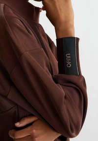 Brązowa kurtka zapinana na zamek z teksturowanej tkaniny. Wyróżniający się czarny mankiet z logo i kontrastowym przeszyciem. Skupienie na detalu rękawa.